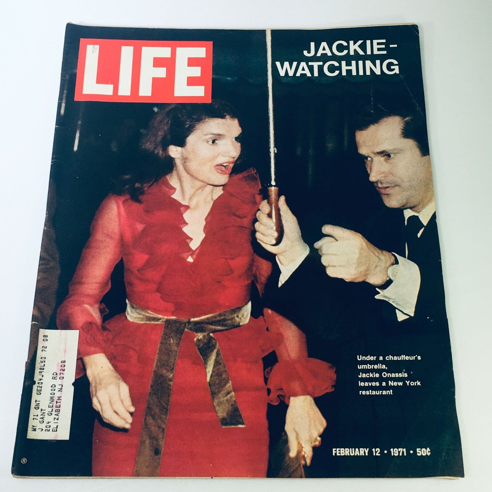 Cover of the February 12, 1971 issue of Life Magazine featuring Jackie Onassis leaving a New York restaurant, dressed in a red ruffled dress, under a chauffeur's umbrella, capturing a moment of glamour and intrigue.
