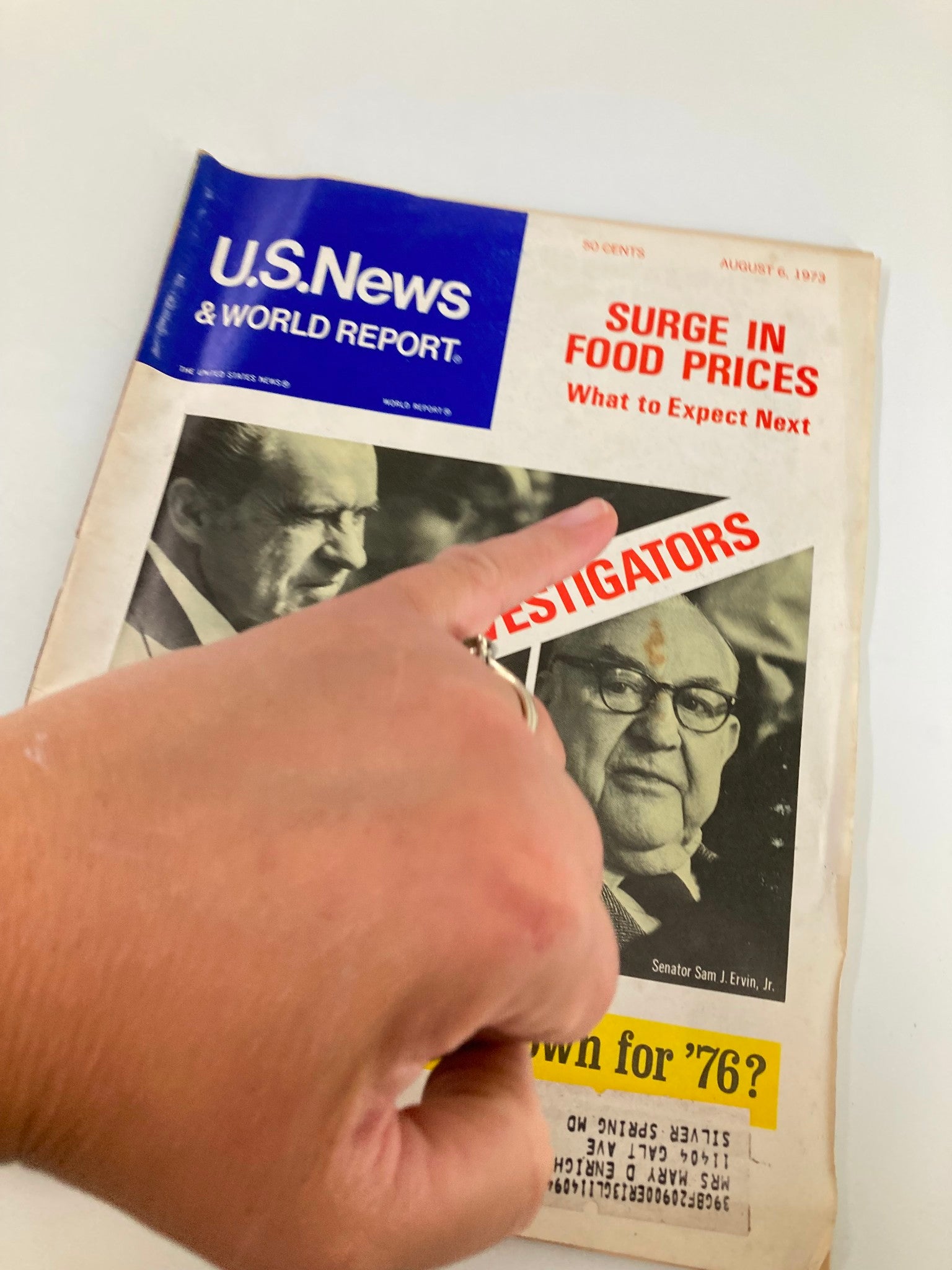 Hand pointing at the cover of US News & World Report Magazine August 6, 1973, which includes headlines about 'Surge in Food Prices' and 'Nixon vs The Investigators'.