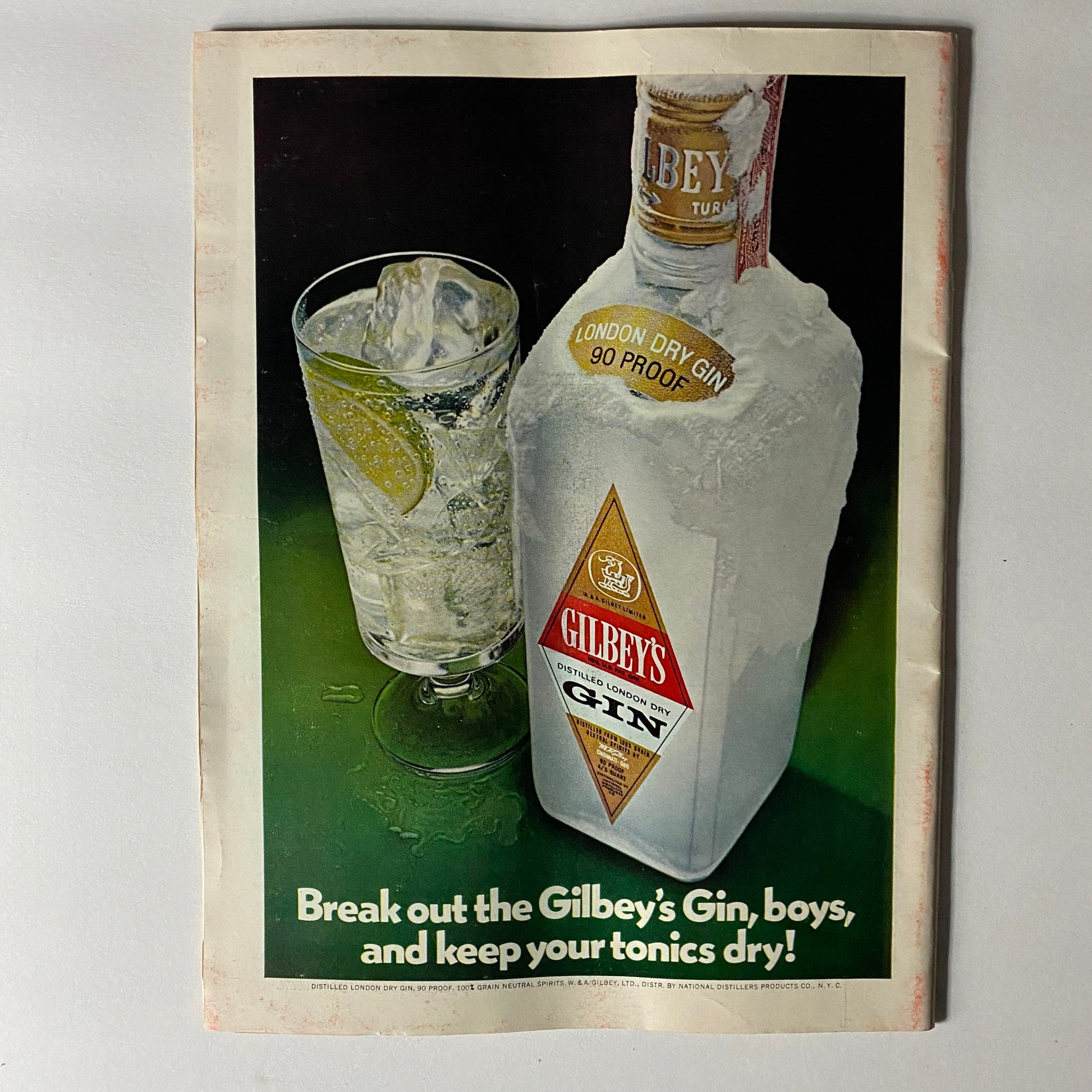 Vintage advertisement for Gilbey's London Dry Gin featuring a bottle and a cocktail glass with ice and lime.