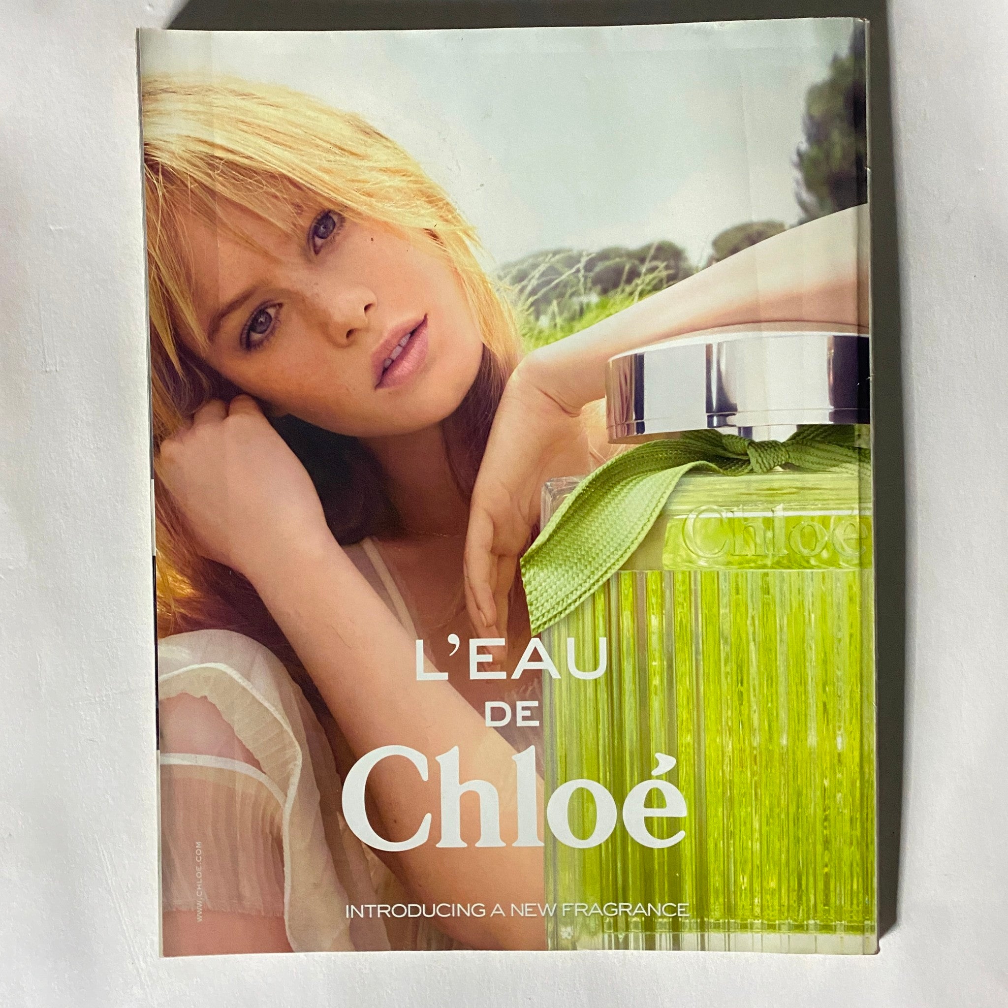 Elle Magazine August 2012 advertisement for L’Eau de Chloé fragrance, featuring a model with blonde hair posing beside the green, stylish perfume bottle, emphasizing the fresh and vibrant essence of the fragrance.