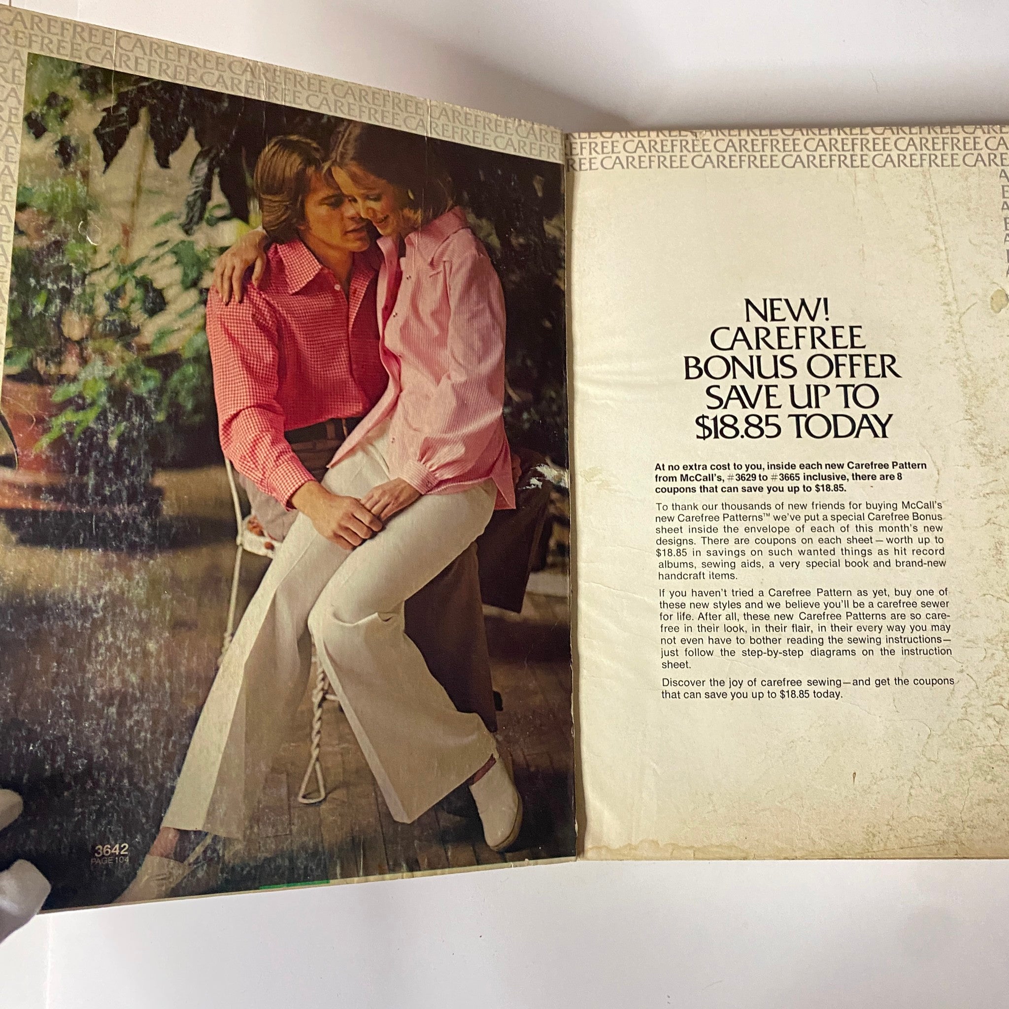 1973 July - August McCall's Carefree Patterns Catalog featuring a couple in stylish 1970s fashion. The male model wears a red checkered shirt, while the female model dons a pink blouse. They are seated in a cozy, nature-inspired setting.
