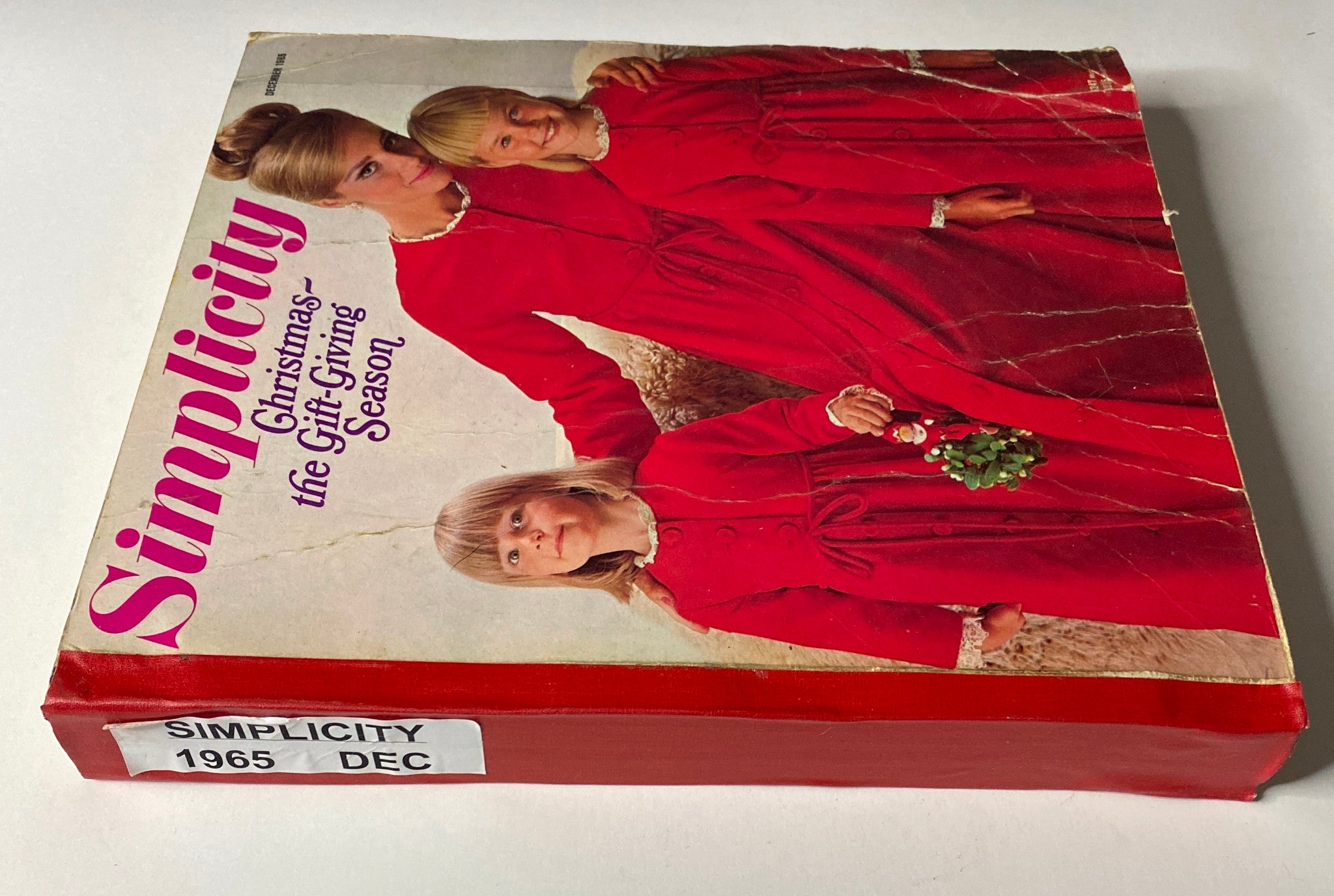 Side view of the 1965 December Simplicity Christmas Catalog, highlighting the cover design with the title and showcasing three figures in red holiday apparel.