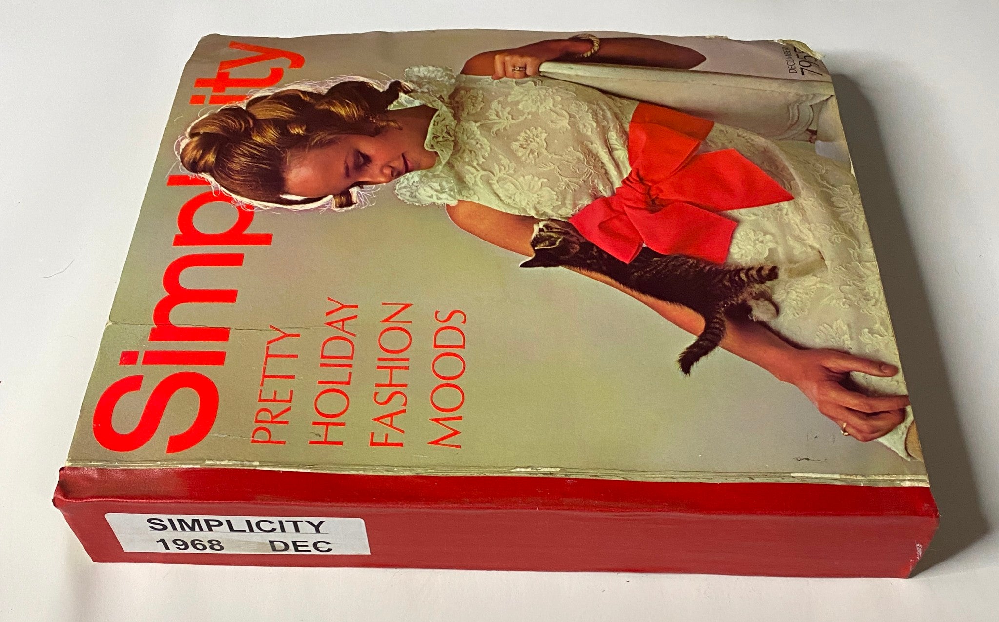 1968 December Simplicity Catalog Sewing Pattern side view showing the distinctive red spine and cover branding, featuring a woman in a stylish dress and kitten, emphasizing holiday fashion.