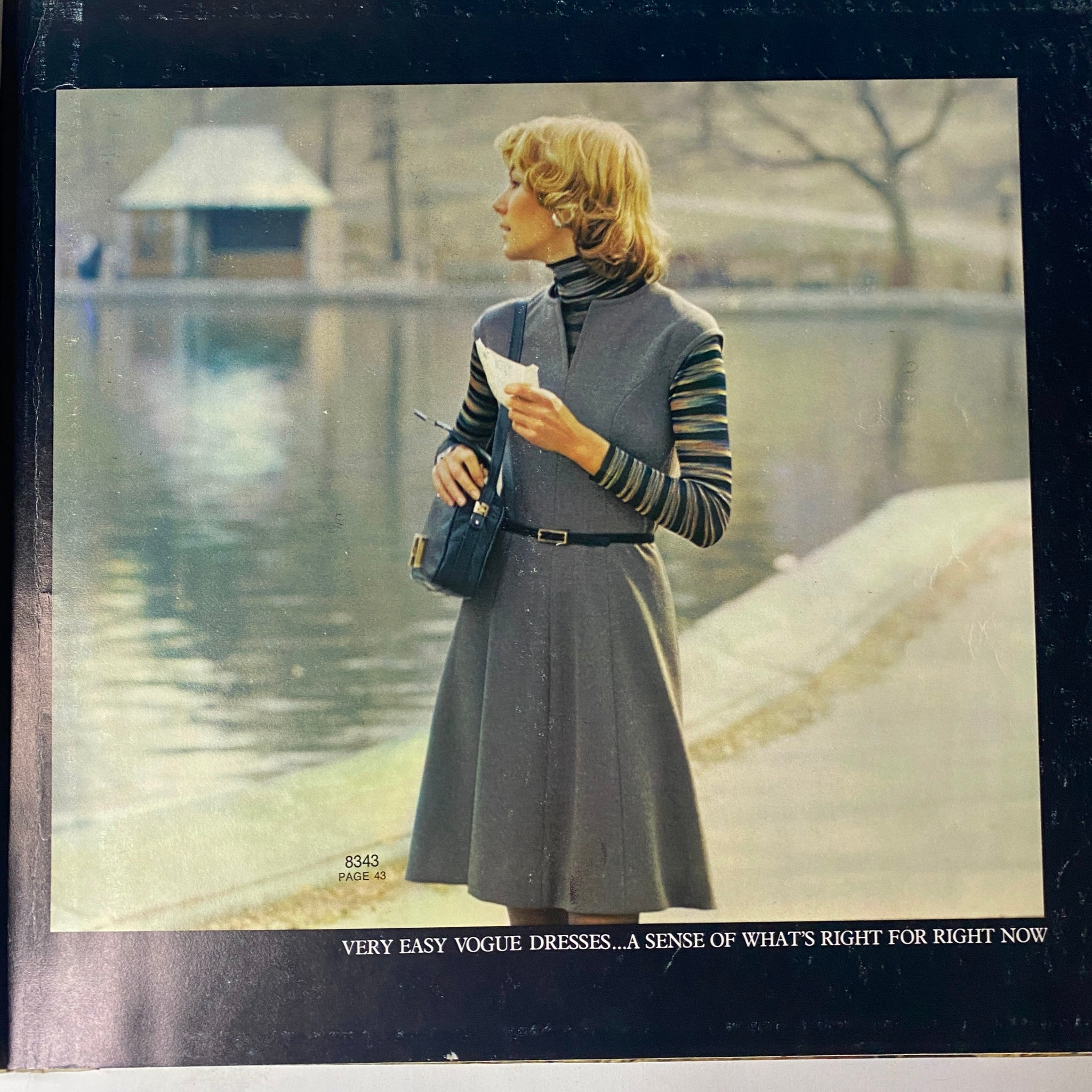 1972 Vogue Patterns September Catalog cover featuring a stylish woman with a chic haircut, wearing a grey sleeveless dress and striped long-sleeve shirt, standing by a serene water body.