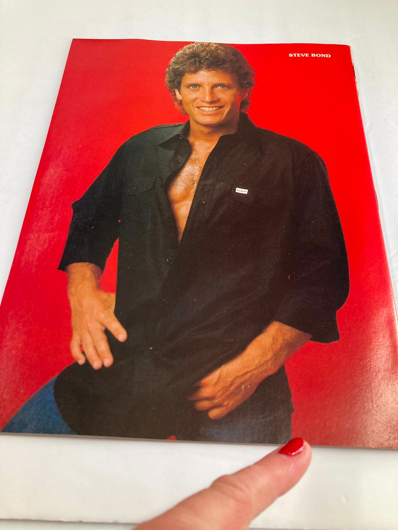 Vintage photo of Steve Bond smiling in the VTG Hollywood's Sexy Rebels Yearbook 1984, dressed in a unbuttoned black shirt, vibrant red background.