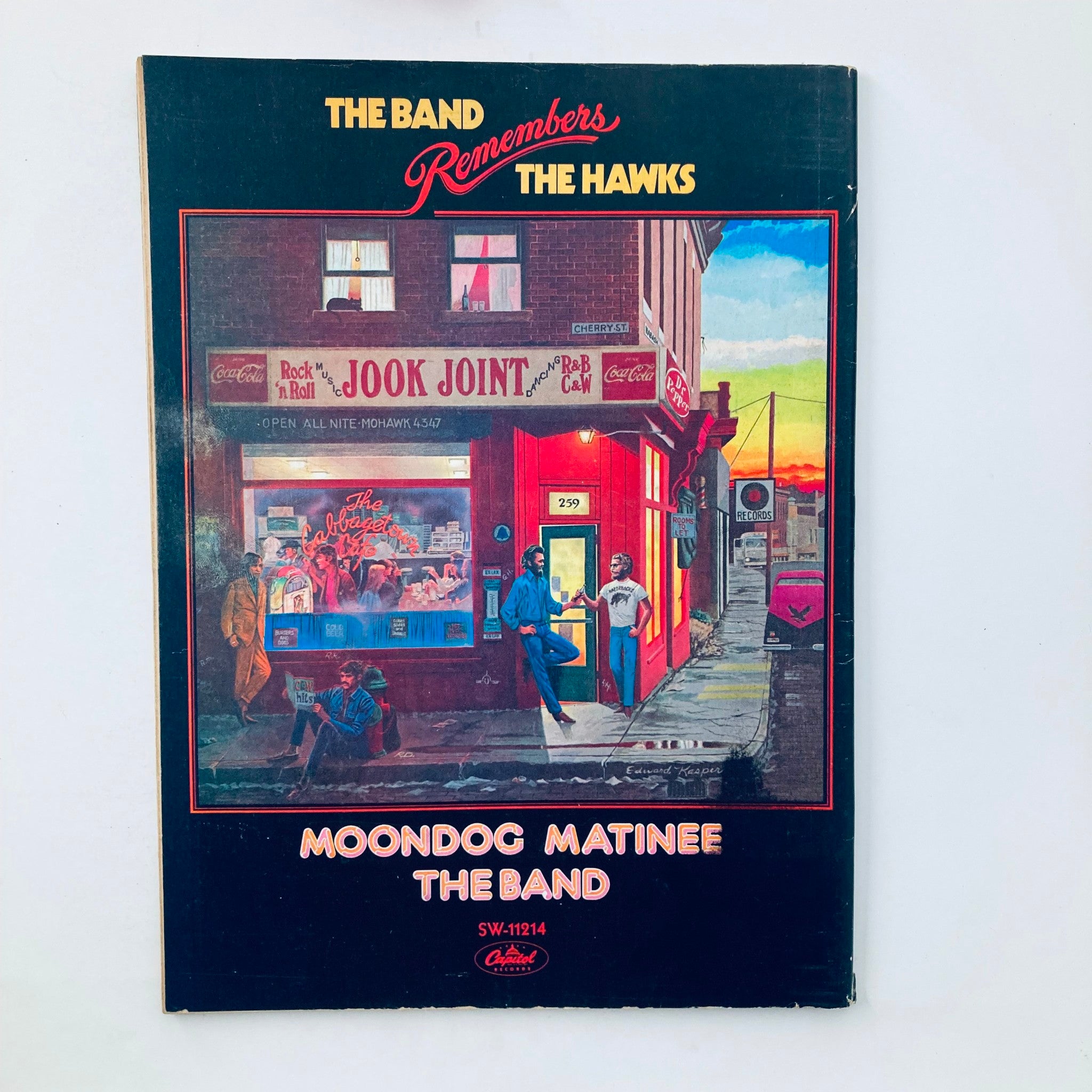 Cover of 'The Band Remembers The Hawks' album, featuring a vibrant illustration of a street scene at sunset with a storefront named 'Jook Joint.' Vintage design captures the essence of classic music.