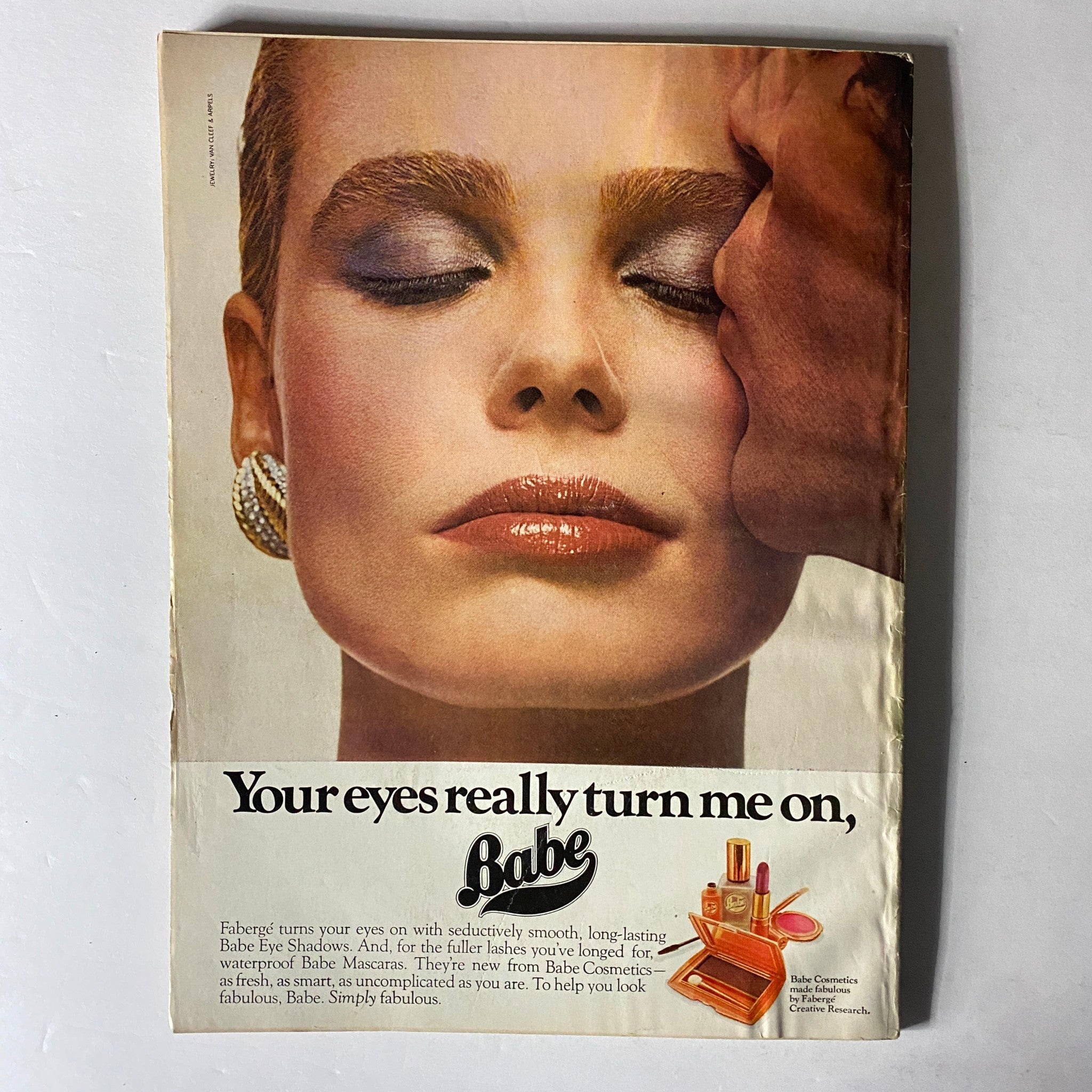 Harper's Bazaar Magazine May 1977 advertisement for Babe Eye Shadows, showcasing a glamorous makeup look with a model's face and a close-up of cosmetic products.