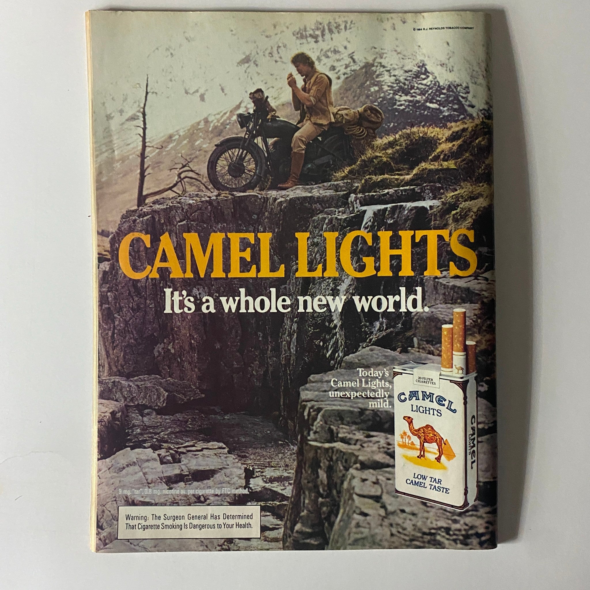 Advertisement from a vintage magazine featuring Camel Lights cigarette, with the slogan 'It's a whole new world' and an image of a man on a motorcycle in a scenic mountainous setting.