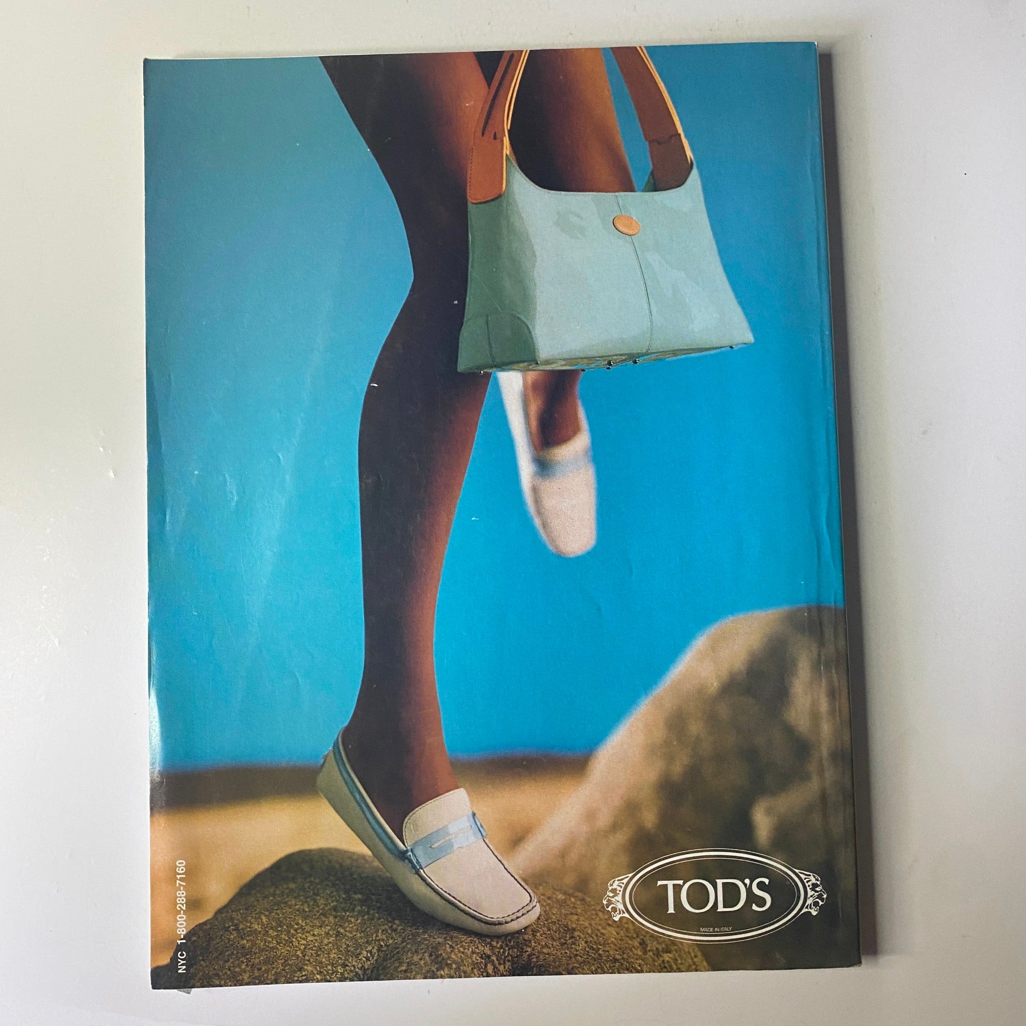 Cover of Tod's magazine featuring a model's legs in boat shoes and a light blue handbag against a beach backdrop. The image emphasizes stylish summer footwear, aligning with the vintage aesthetic of the brand.