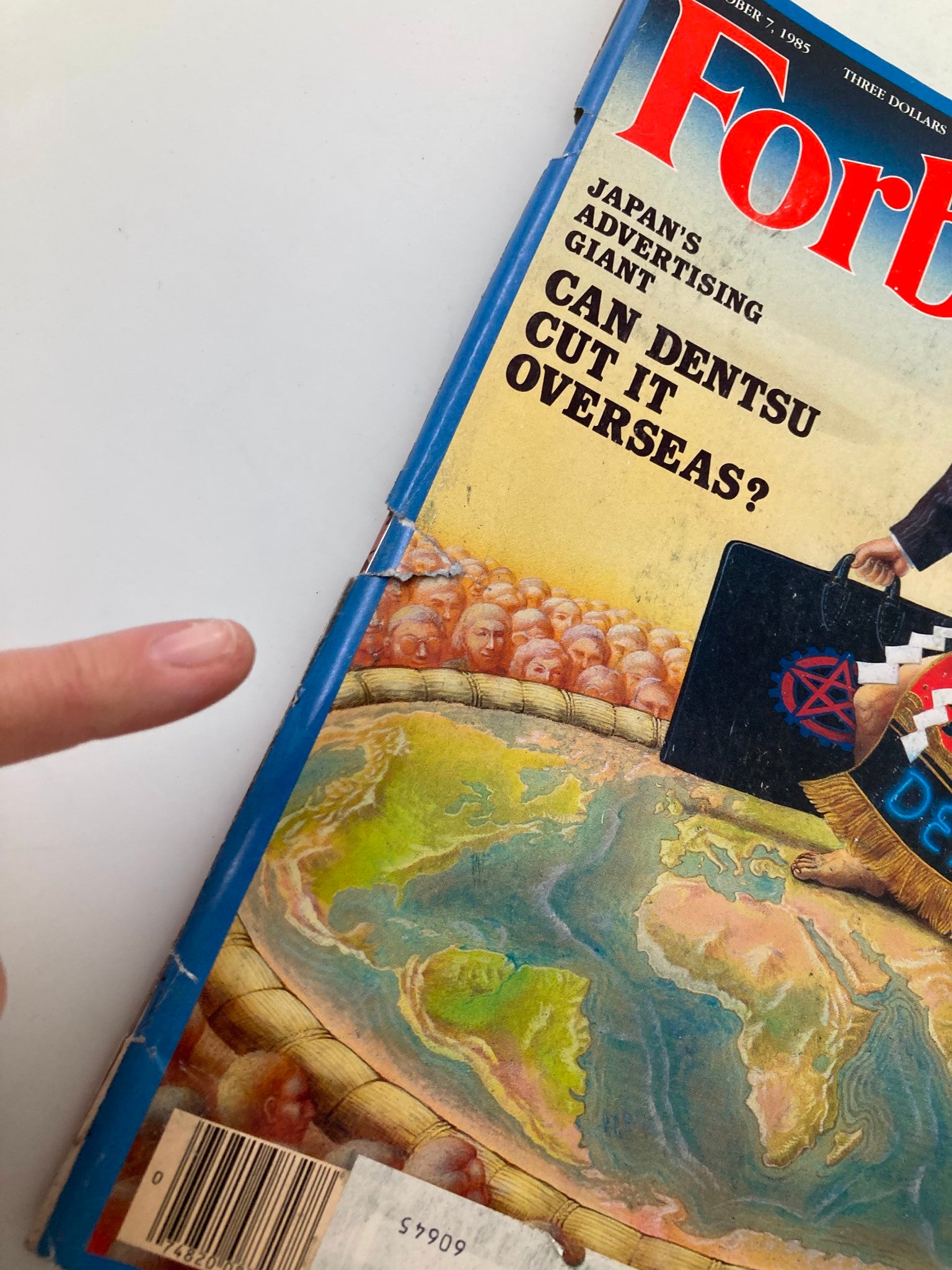 Close-up of the spine of the vintage Forbes Magazine from October 7, 1985, highlighting the title 'Japan's Ad Giant: Can Dentsu Cut It Overseas?' with visible wear and a finger pointing at the pages.
