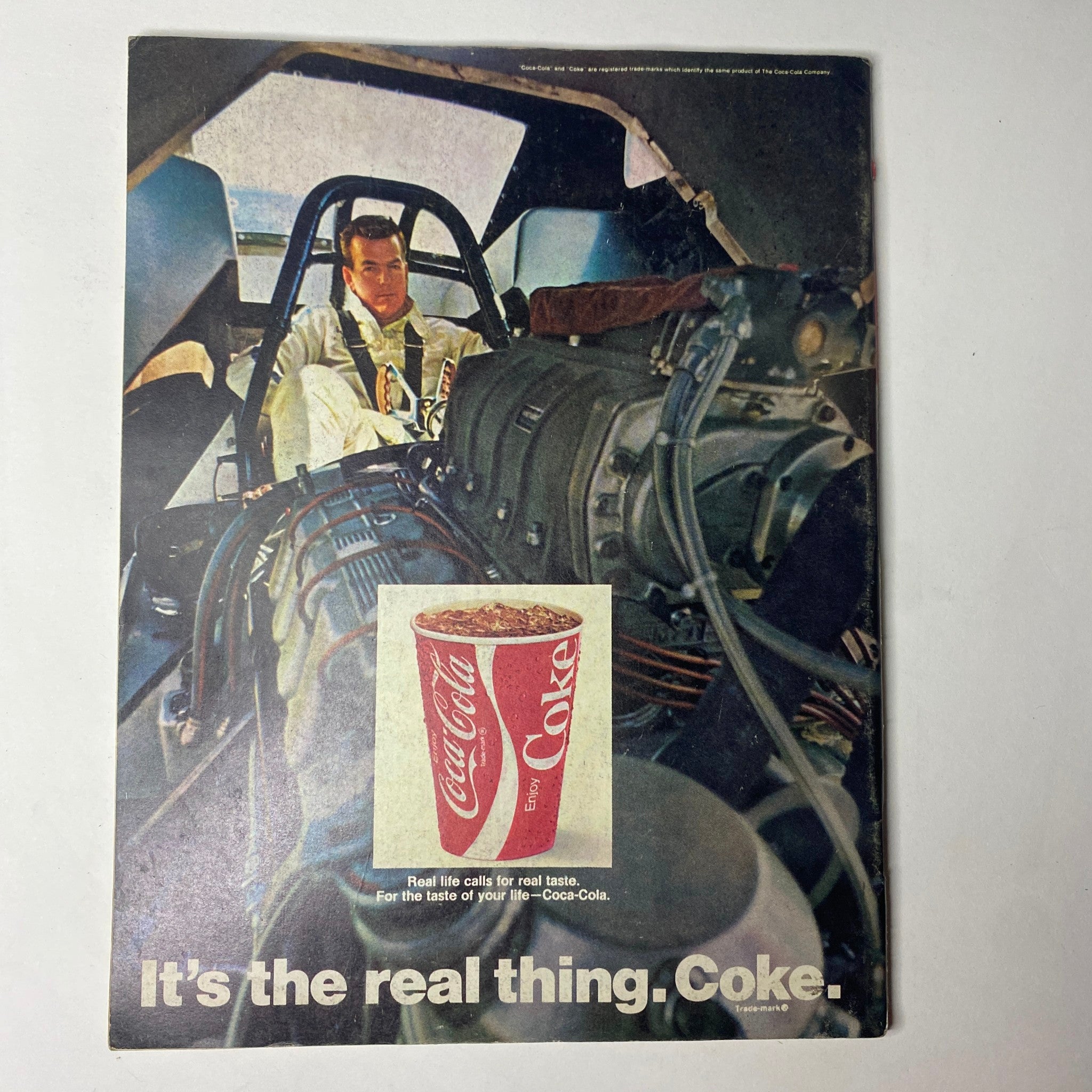 Promotional image for Coca-Cola featuring a driver in a race car, emphasizing the slogan 'It's the real thing', as seen in Car Craft Magazine June 1970 Vol 18 No. 6 Supercar Swan Song.