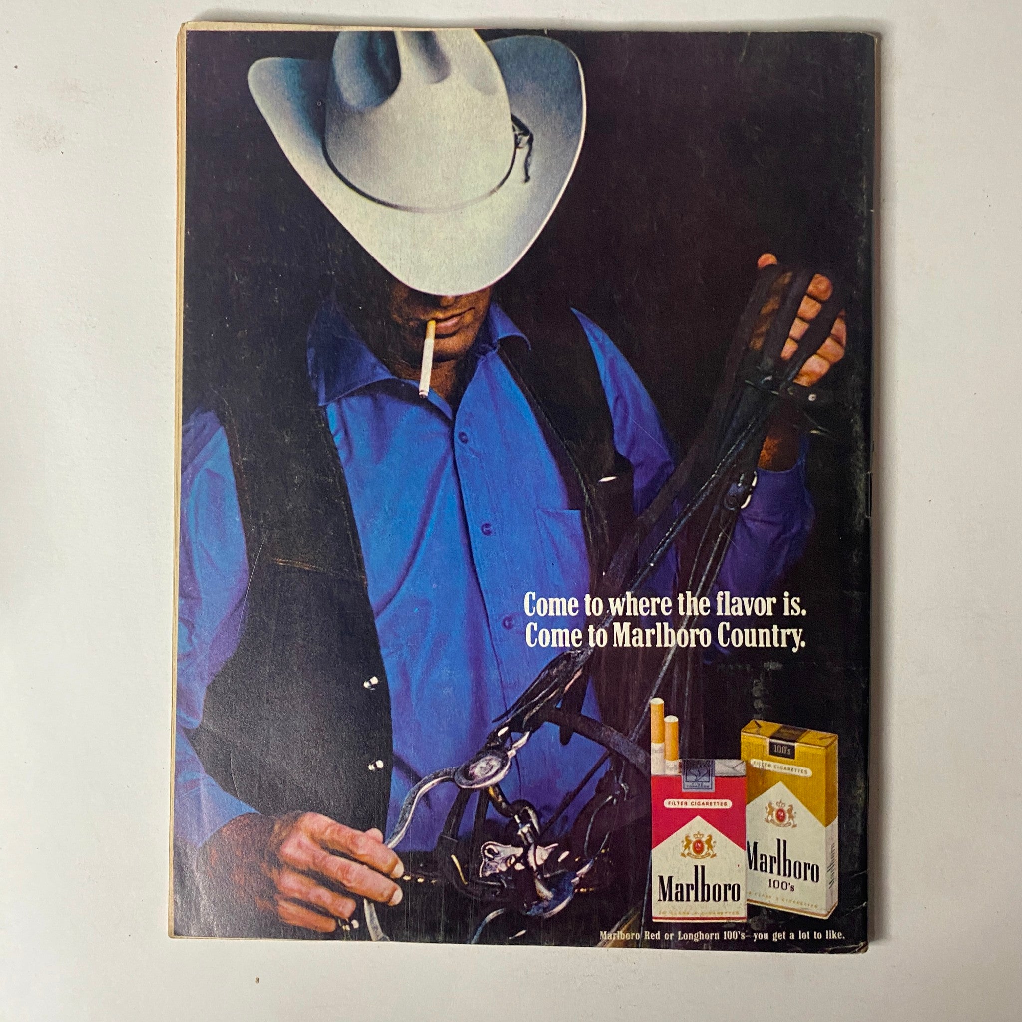Vintage Marlboro advertisement featuring a man in a cowboy hat holding a saddle, promoting the tagline 'Come to where the flavor is. Come to Marlboro Country.' Related to the Car Craft Magazine April 1970 issue, showcasing the era's strong connections to Americana and lifestyle branding.