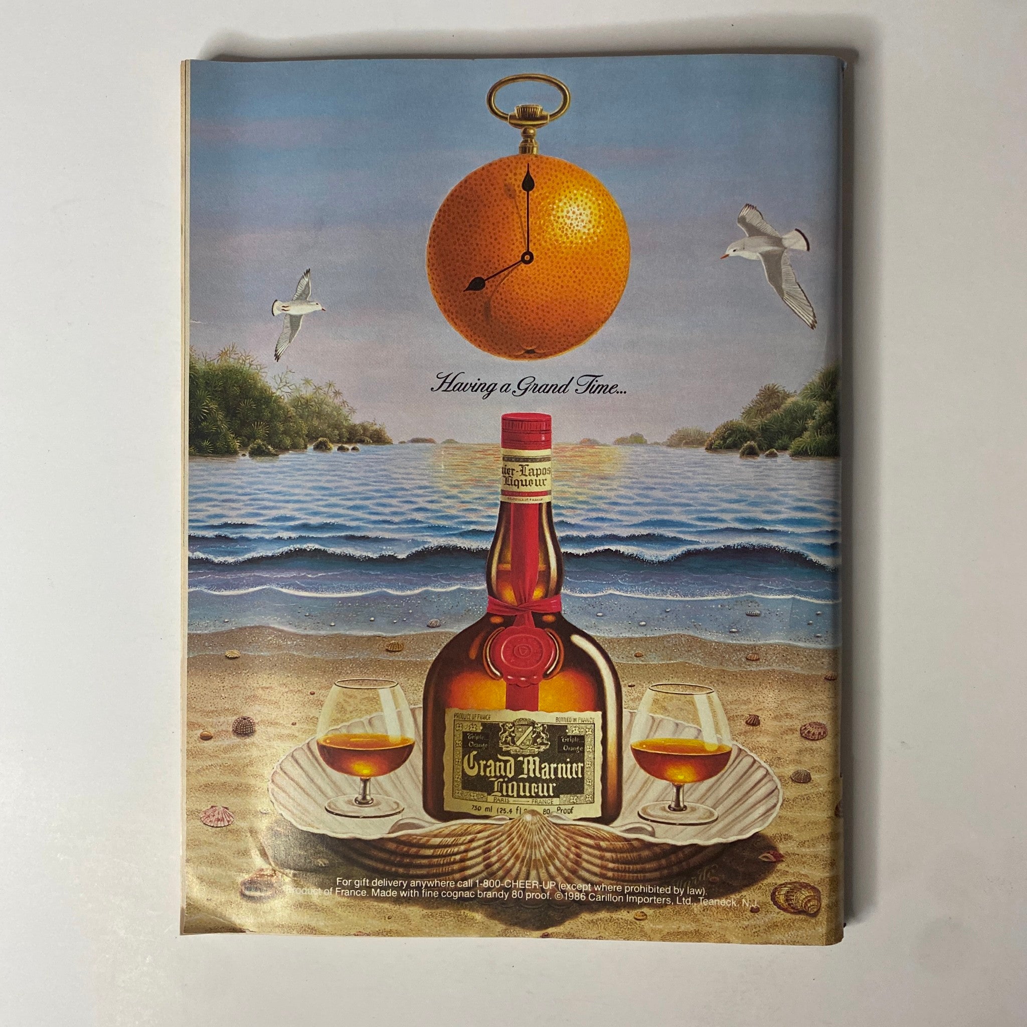 Colorful advertisement for Grand Marnier from Forbes Magazine September 8, 1986. Features a whimsical illustration of a clock and orange over a beach setting, promoting the liqueur. Ideal for vintage magazine enthusiasts and advertising collectors.