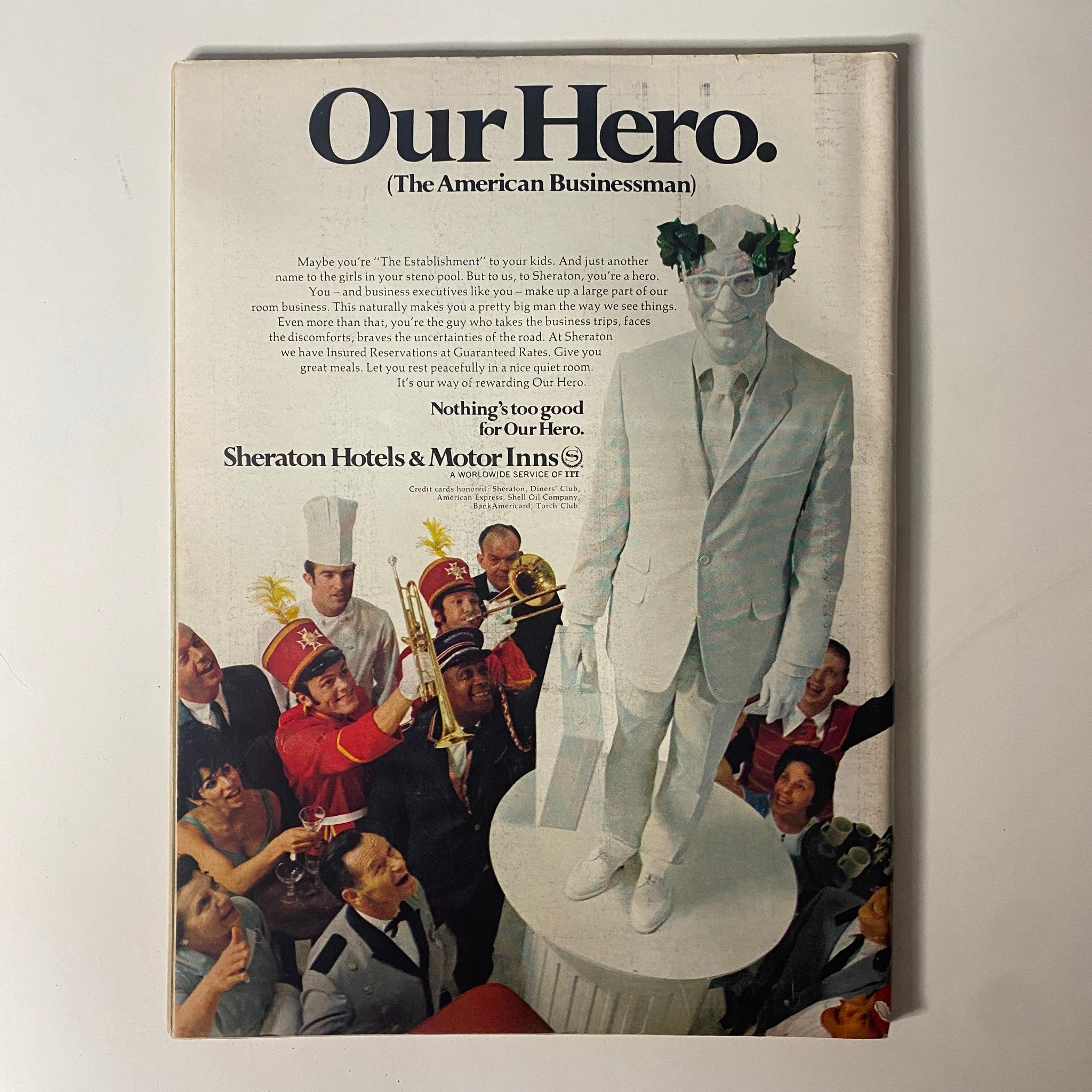 Advertisement page from a magazine featuring a character in a white suit symbolizing 'Our Hero' (The American Businessman), surrounded by celebratory figures, representing the themes of corporate success.