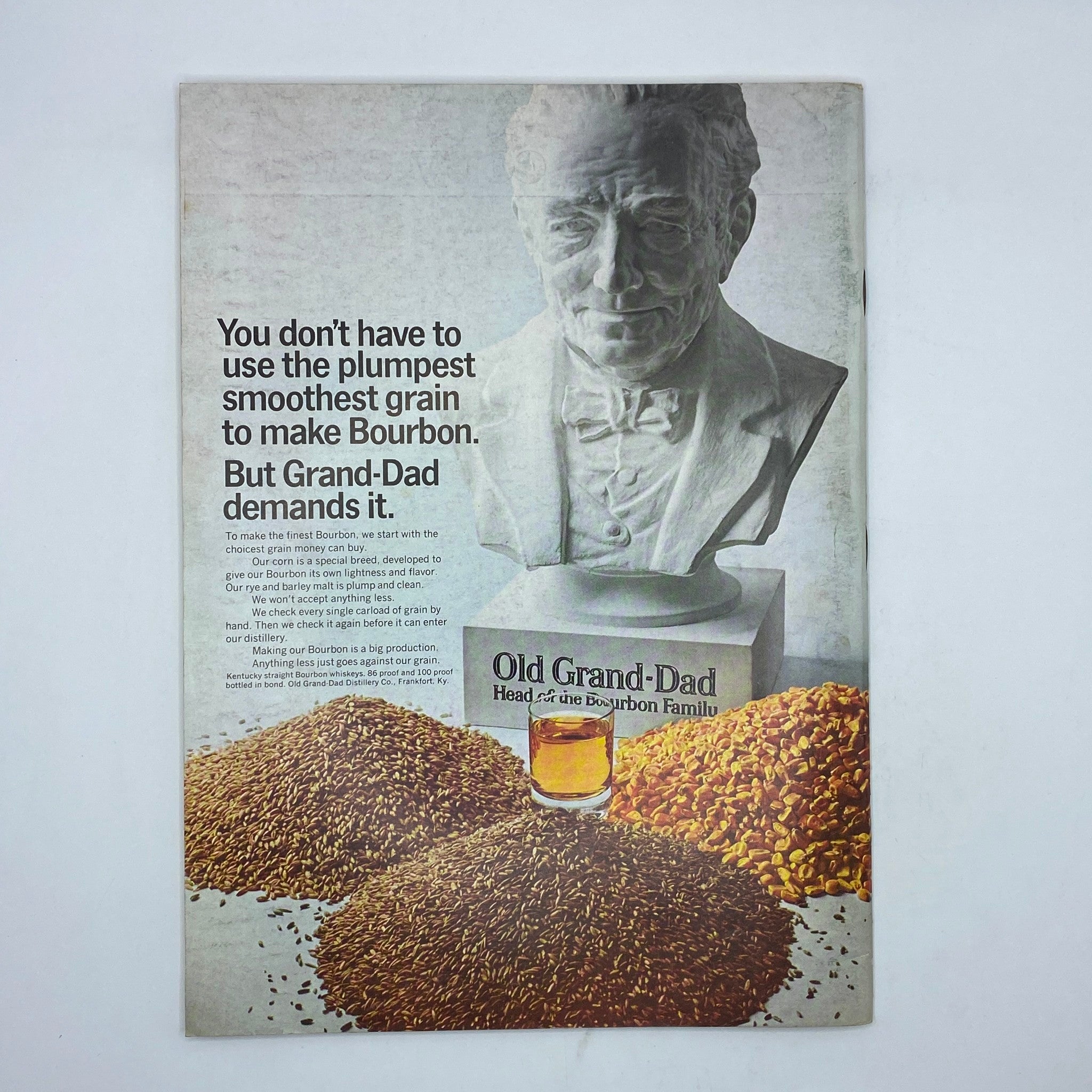 Old Grand-Dad Bourbon advertisement featuring a statue of a distinguished gentleman, with grains and whiskey glass at the forefront. The ad states: 'You don’t have to use the plumpest smoothest grain to make Bourbon. But Grand-Dad demands it.' Captured in a vintage style, enhancing the quality of craftsmanship associated with Old Grand-Dad bourbon.