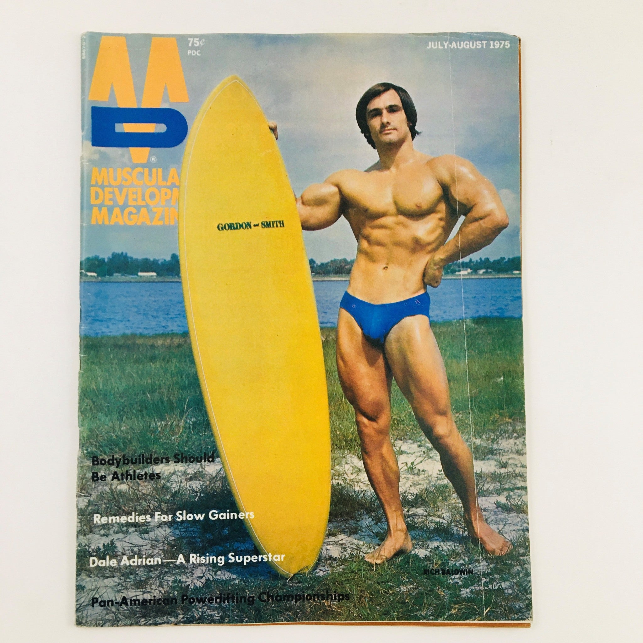 Muscular Development Magazine July 1975 featuring bodybuilder Rich Baldwin on the cover, posing with a yellow surfboard by a lakeside backdrop. This vintage bodybuilding magazine showcases fitness culture of the 1970s.