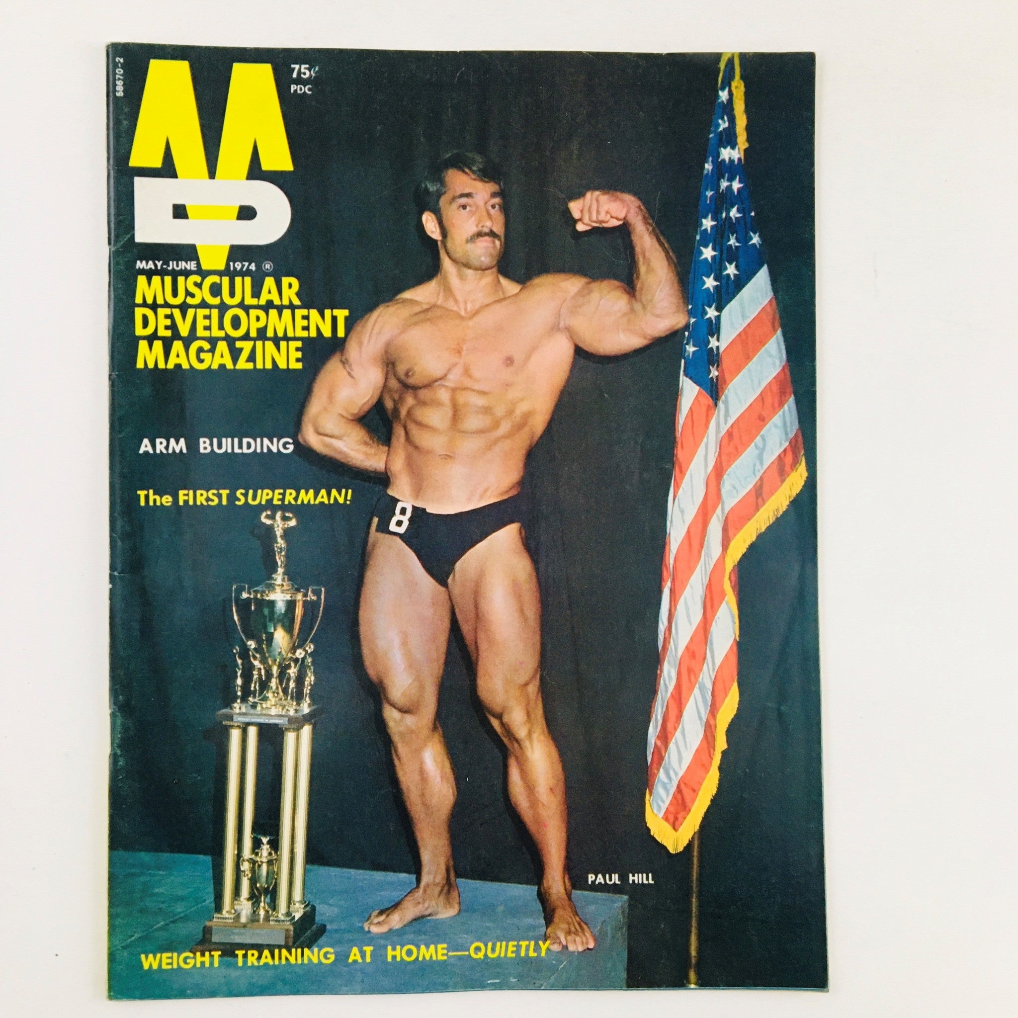 Cover of Muscular Development Magazine May 1974 featuring Paul Hill, The First Superman, showcasing arm building with a trophy and an American flag.