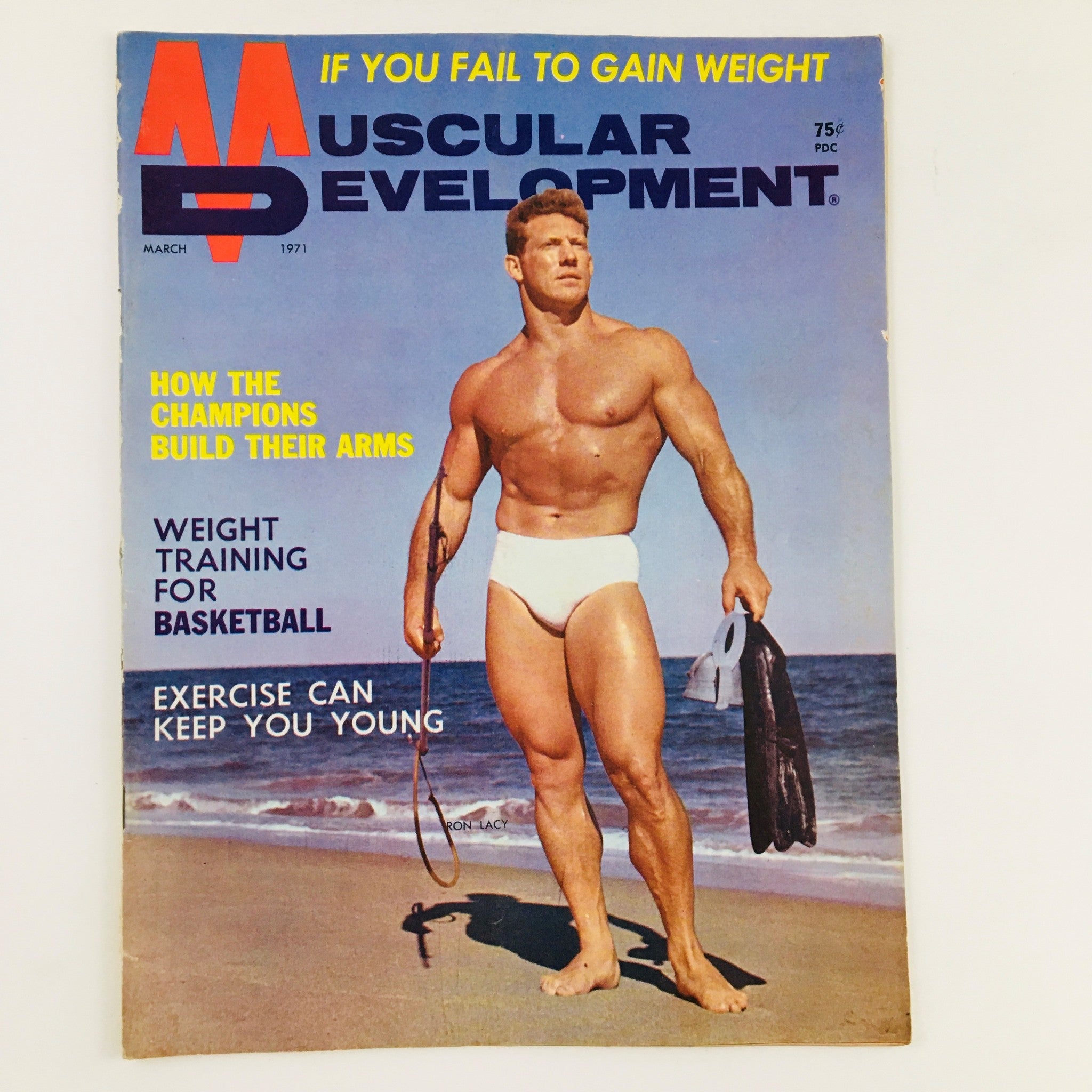Cover of Muscular Development Magazine March 1971 featuring Ron Lacy, a shirtless bodybuilder posing on the beach with a swim fin in hand, highlighting the themes of weight training and fitness.