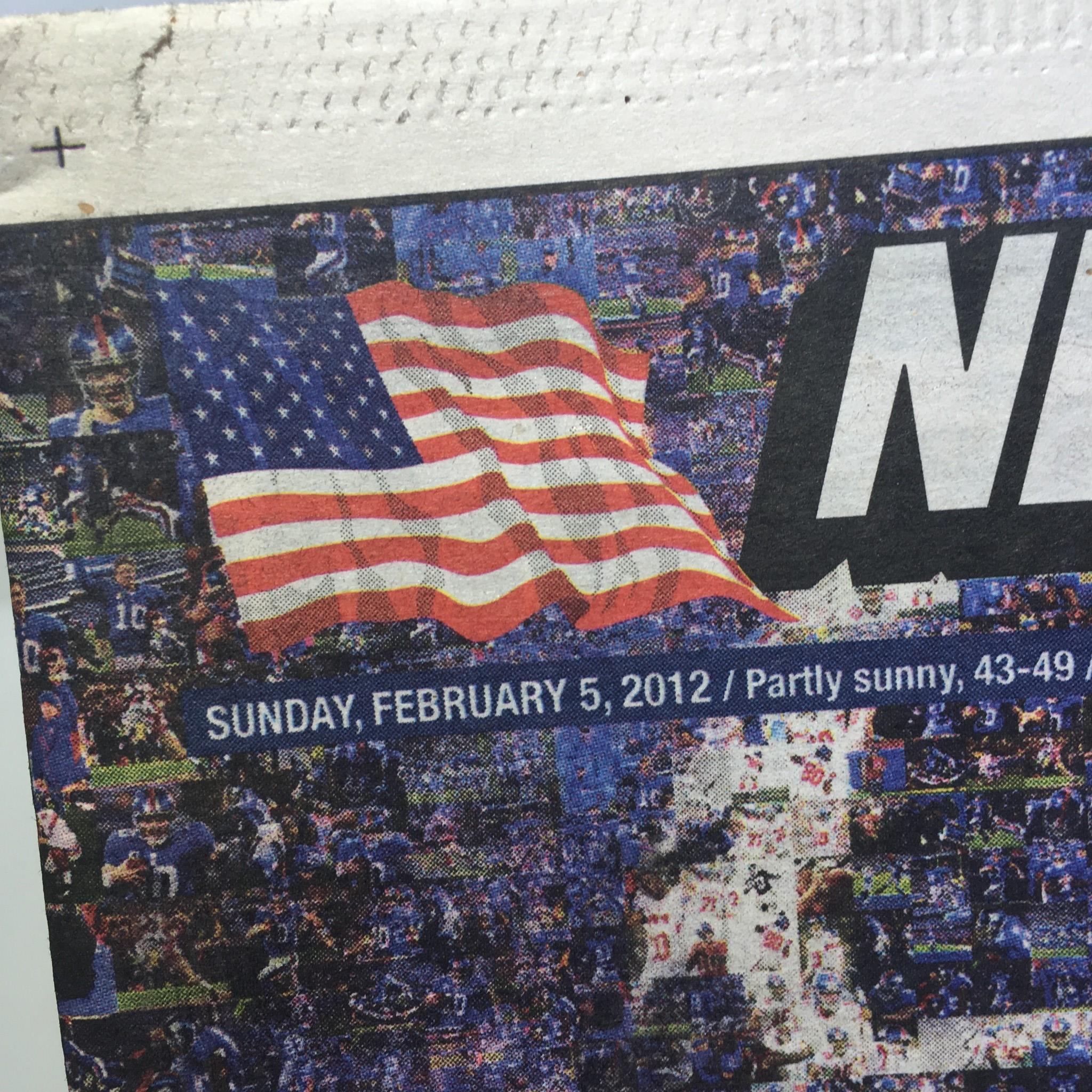 Close-up of the New York Post dated February 5, 2012, highlighting a faded American flag in the corner and the date. The newspaper includes the title 'Standing on the Shoulders of Giants' and reflects a special Super Bowl edition.