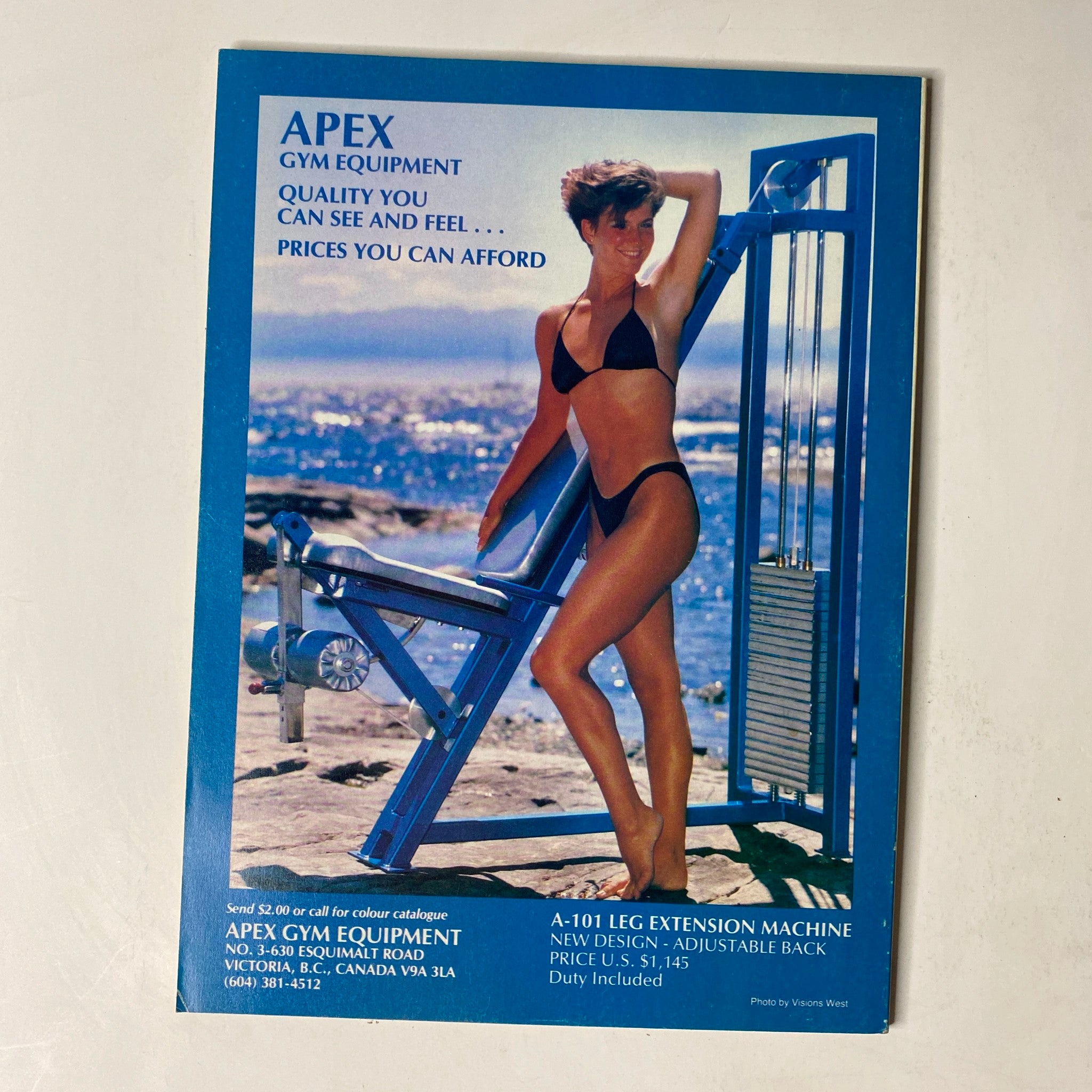 Advertisement for Apex Gym Equipment in MuscleMag International Magazine October 1989, featuring a woman in a bikini demonstrating the adjustable leg extension machine with a view of the ocean.